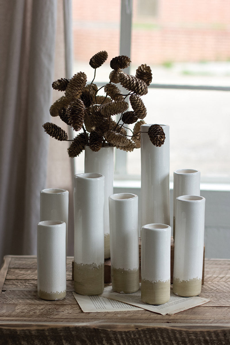 several sizes of cylinder shaped vase arranged together on a wooden table in front of a window. one vase has a cluster of small pinecones in it.