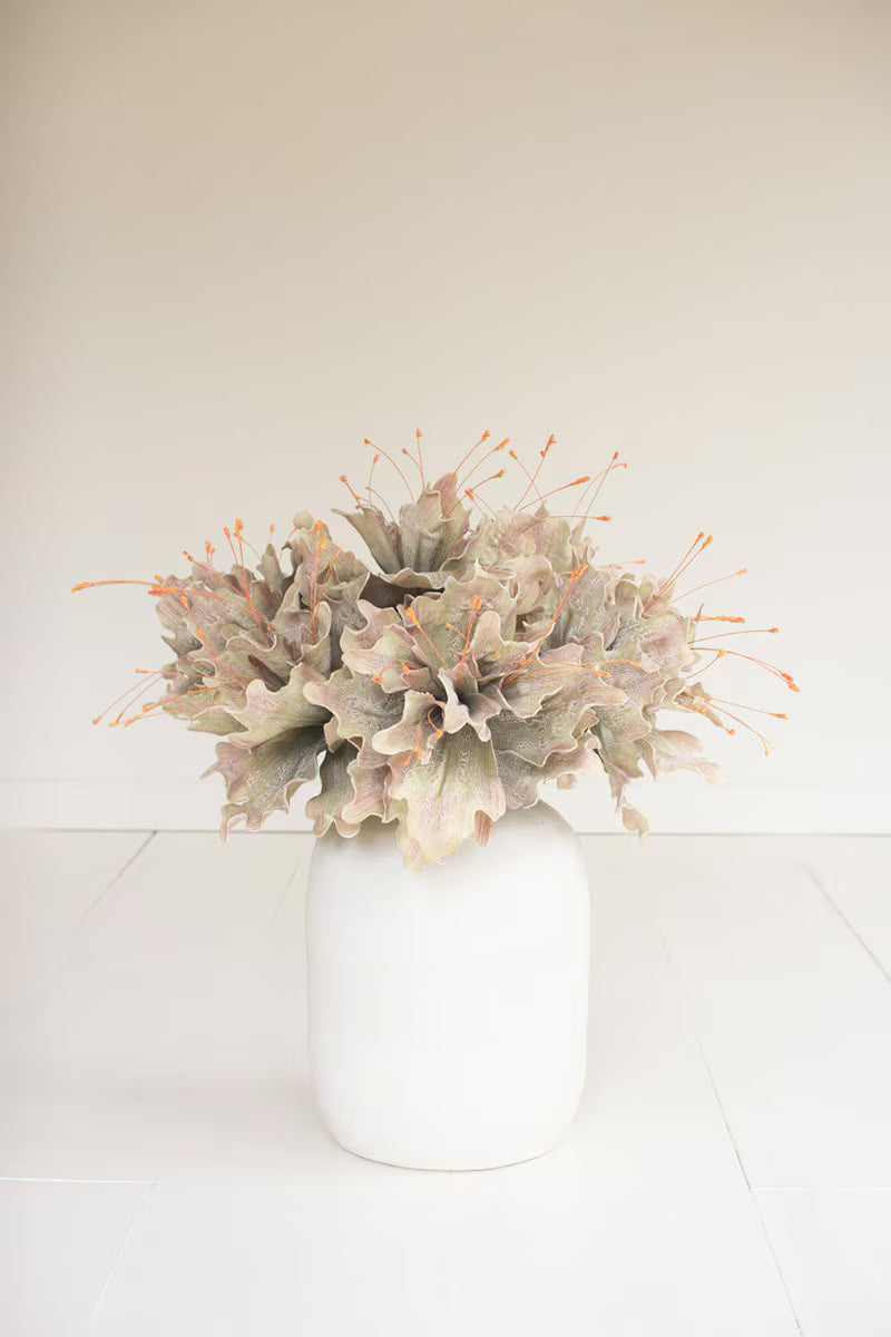 A floral arrangement featuring dusty yellow green and muted pink leaves with orange-tipped spindles, presented in a matte white ceramic vase against a clean, off-white background.
