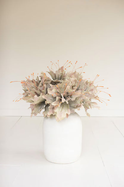 A floral arrangement featuring dusty yellow green and muted pink leaves with orange-tipped spindles, presented in a matte white ceramic vase against a clean, off-white background.