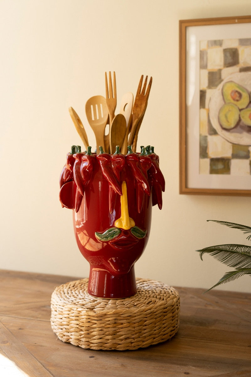 Red ceramic head vase with wooden utensils in it on a woven base against a neutral wall.
