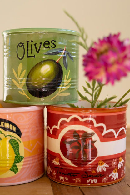 Three decorative ceramic vessels designed to resemble canned goods with illustrations of olives, lemons, or tomatoes stacked on a wooden surface.