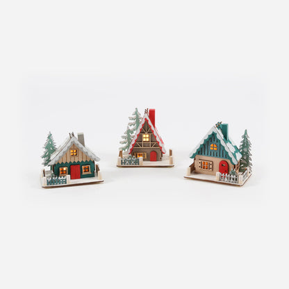 three decorative ski lodge ornaments with different colors and designs, each featuring a small tree and lightsin the windows.