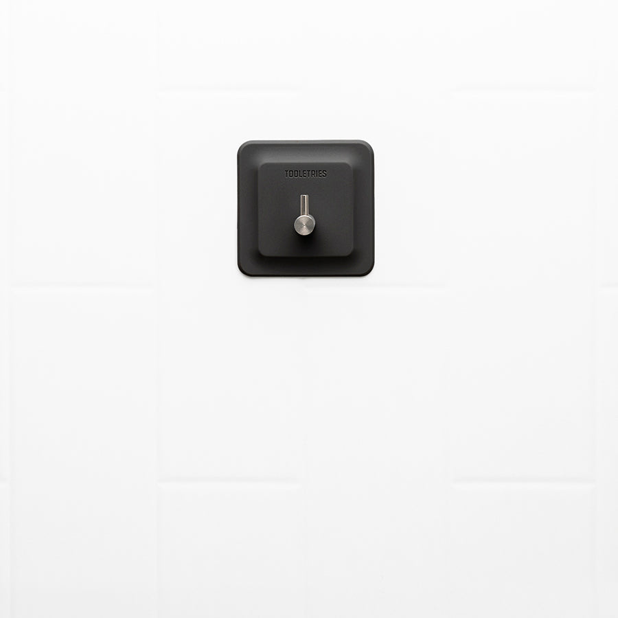 Black silicone square with metal L hook on it, stuck on a white tiled wall.