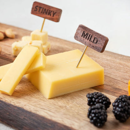 blocks of cheese on a wood board with chees markers in them and blackberries scatter around.