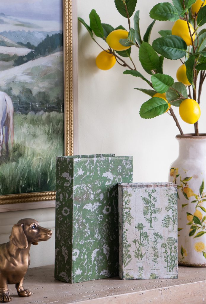 Decorative book boxes with floral patterns on a shelf next to a vase with lemons and a painting.