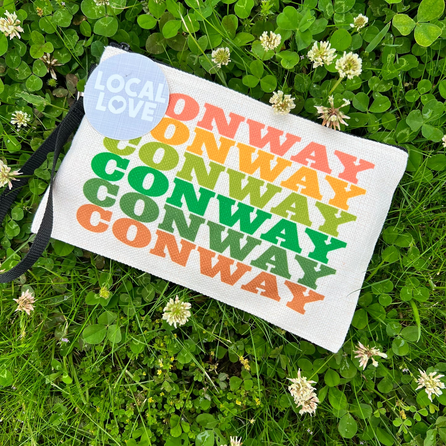 off-white zipper bag with "conway" printed in assorted colors laying in a field of clovers.