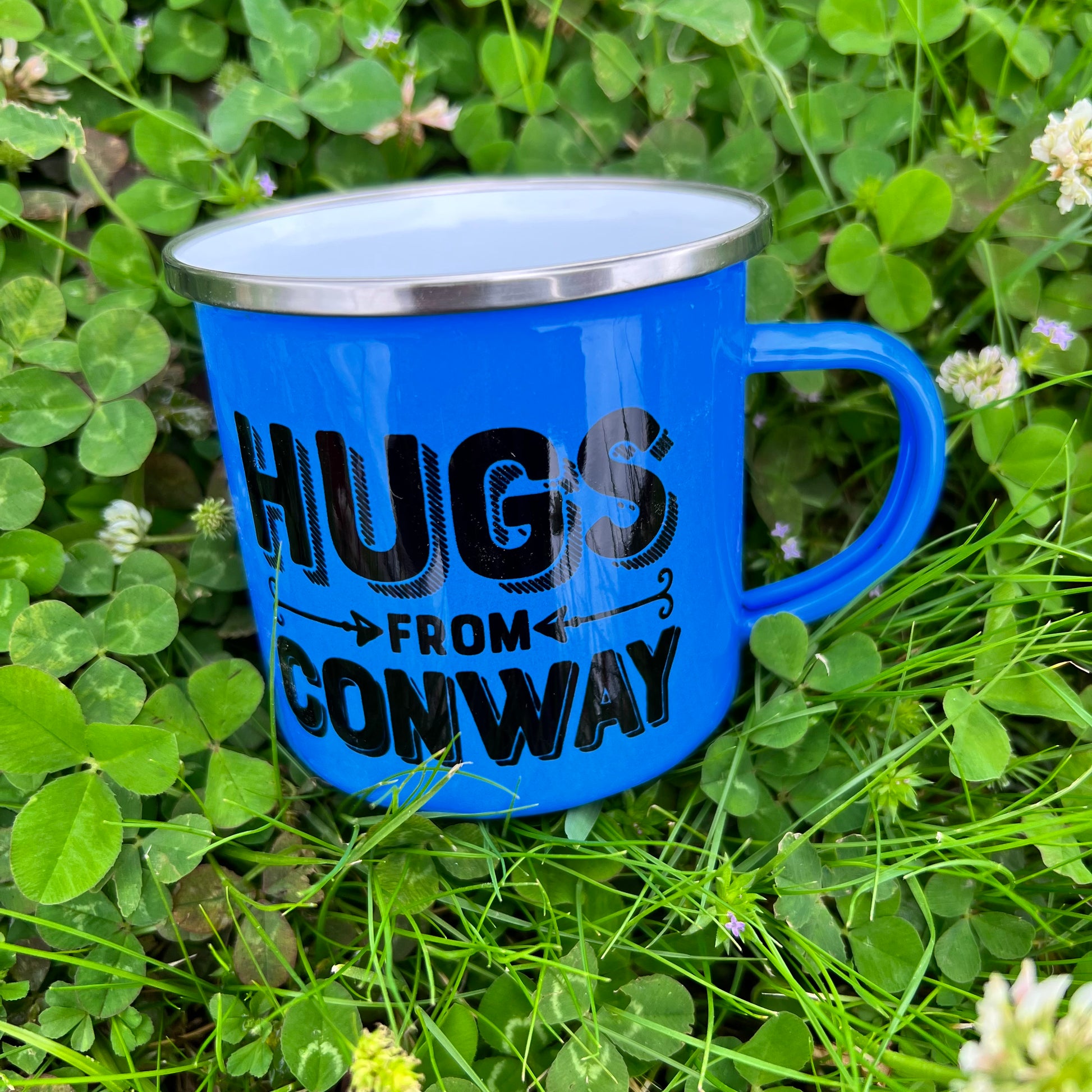 blue  tin mug with "hugs from conway" in black laying in a field of clovers.