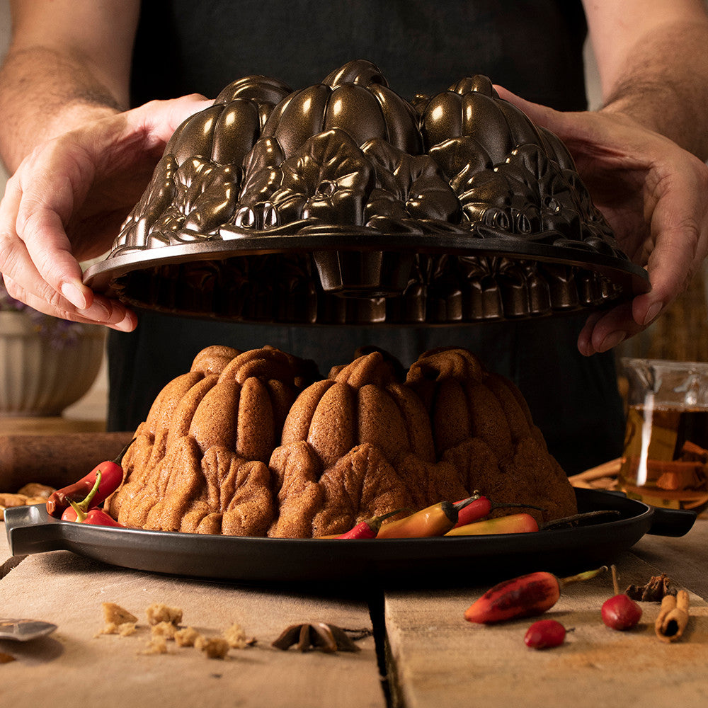 person lifting pumpkin patch bundt pan off of freshly baked cake on a platter on a wooden table.