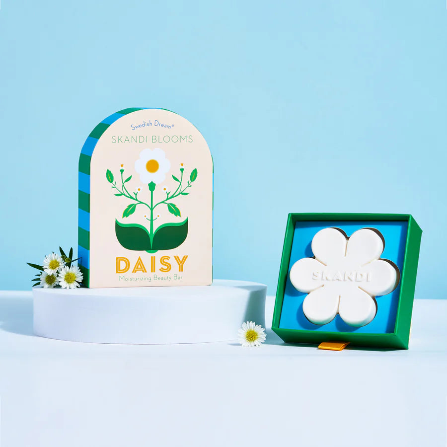 Flower shaped bar of soap in a box set next to its floral packaging with daisies scattered about.