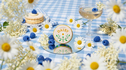 tin of daisy lip balm set on a blue gingham picnic blanket with flowers and berries
