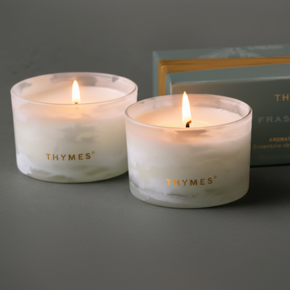 Two lit THYMES Fraser Fin scented candles in clear glass holders with white wax, shown in front of a muted green product box with gold lettering on a dark gray surface.