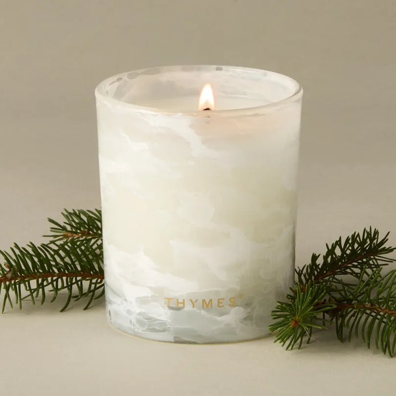 A lit white scented candle in a frosted glass jar with the brand name 'THYMES' printed in gold near the base. The candle is styled with green pine branches on a neutral beige background.