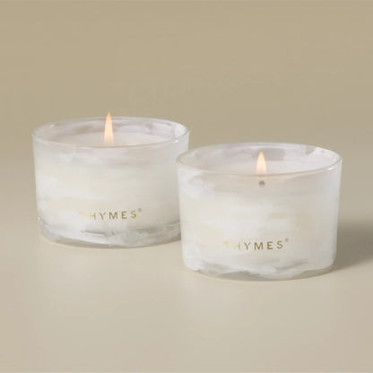 Two lit white candles in clear cylindrical glass holders with gold 'THYMES' branding, set against a warm beige background.