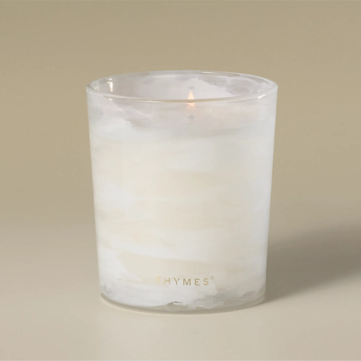A lit white wax candle in a frosted glass jar with the brand name 'THYMES' printed in gold near the bottom, set against a solid beige background.