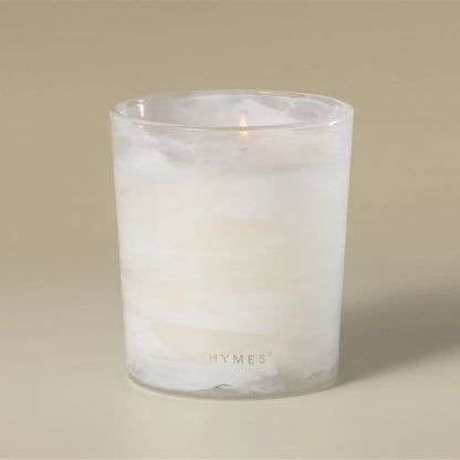 A lit white wax candle in a frosted glass jar with the brand name 'THYMES' printed in gold near the bottom, set against a solid beige background.