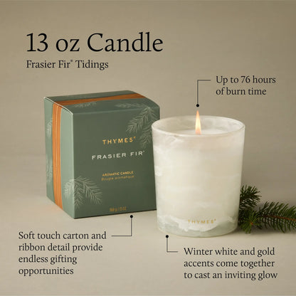 A 13 oz candle by Thymes in the Frasier Fir scent, shown as a lit white wax candle in a glass vessel next to its green and gold gift box, with decorative pine branches on a neutral beige background with descriptive text