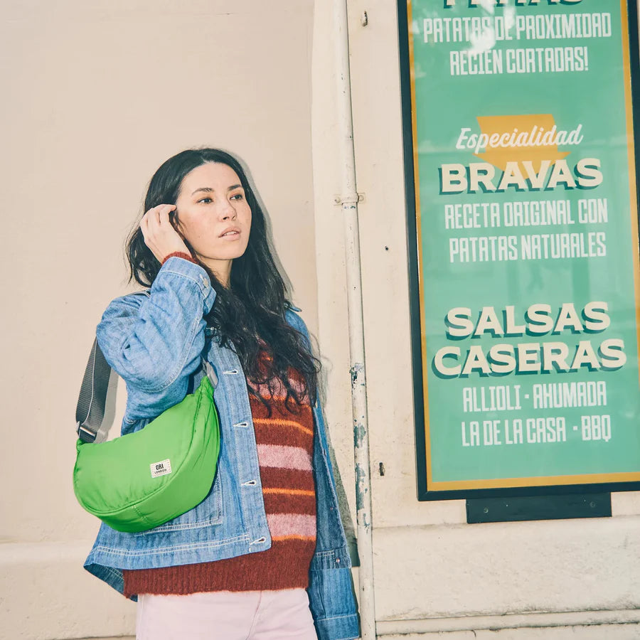 person wearing green crescent shaped shoulder bag with light grey handle on their shoulder and standing next to a restaurant sign