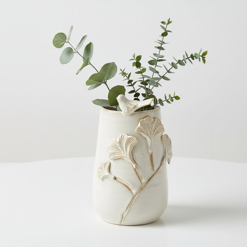 White ceramic vase with leaves and bird on it and greenery in it on a light background