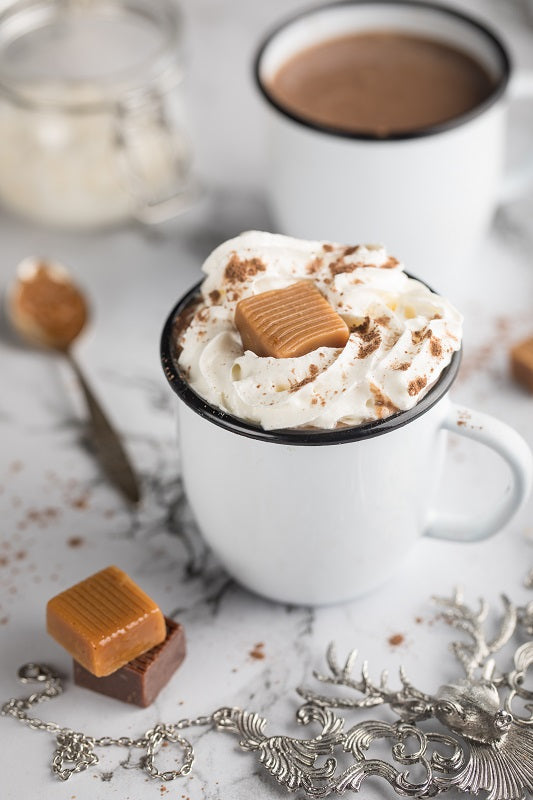 white mug filled with salted caramel hot chocolate set on a table with caramel squares, and holiday decor.