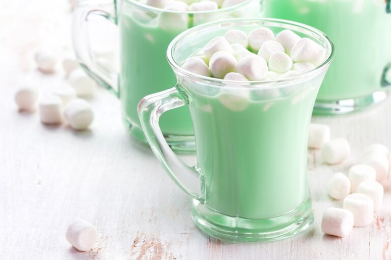 glass mugs filled with dino hot chocolate set on a table with marshmallows scattered around.