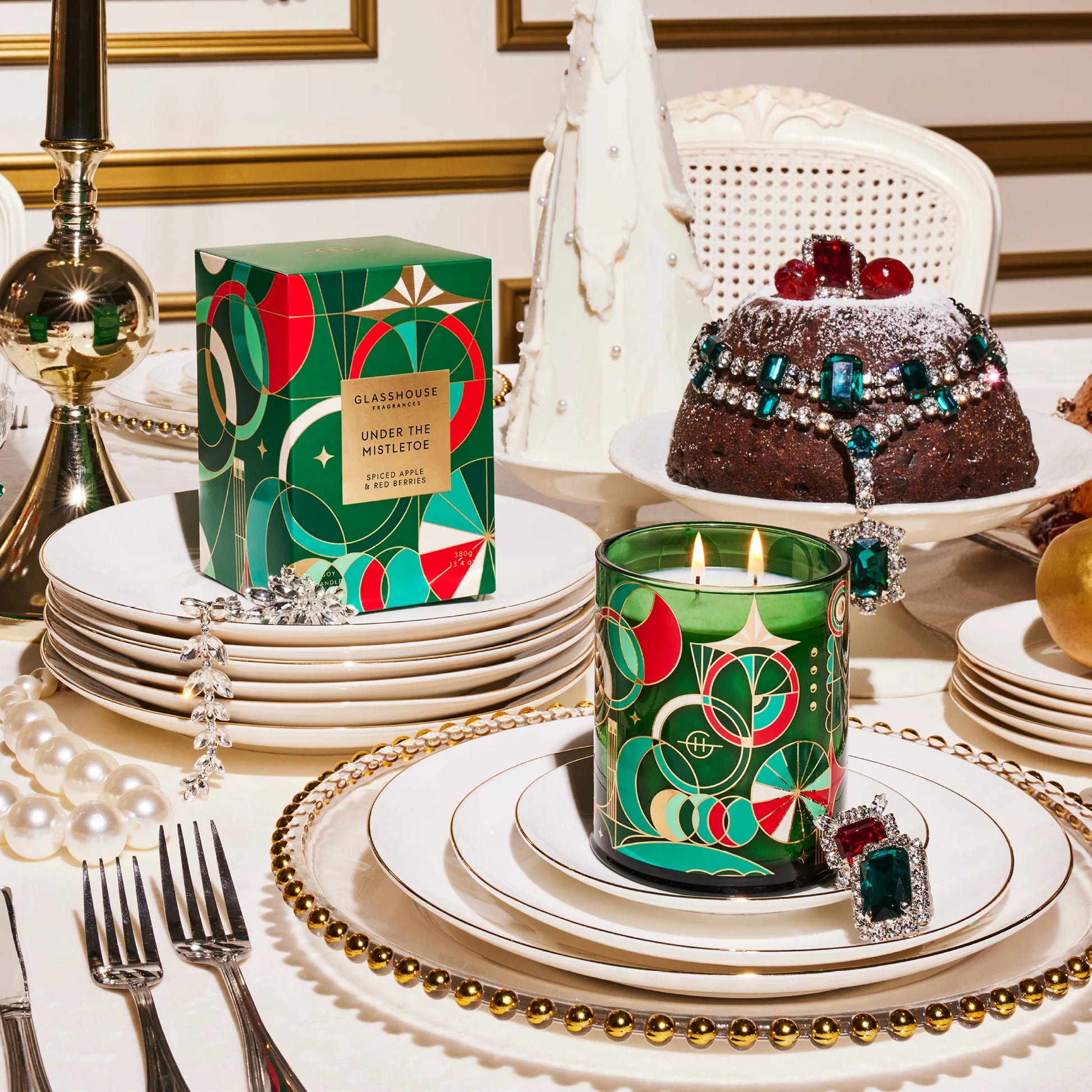 Table set with stacks of dishes and silverware and a cake in the background with a lit under the mistletoe candle on the dishes.