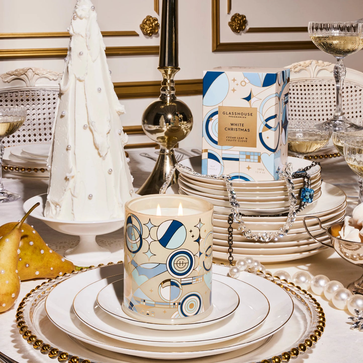 Table set with stacks of dishes and glasses of champagne and jewels draped on them with a lit white Christmas candle on top of a plate.