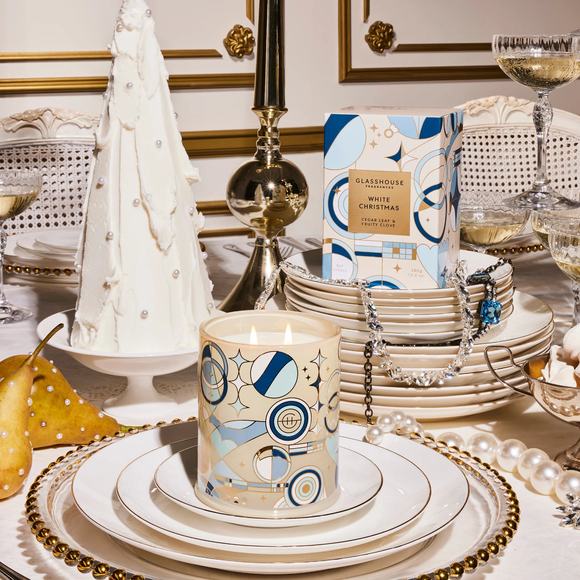 Table set with stacks of dishes and glasses of champagne and jewels draped on them with a lit white Christmas candle on top of a plate.