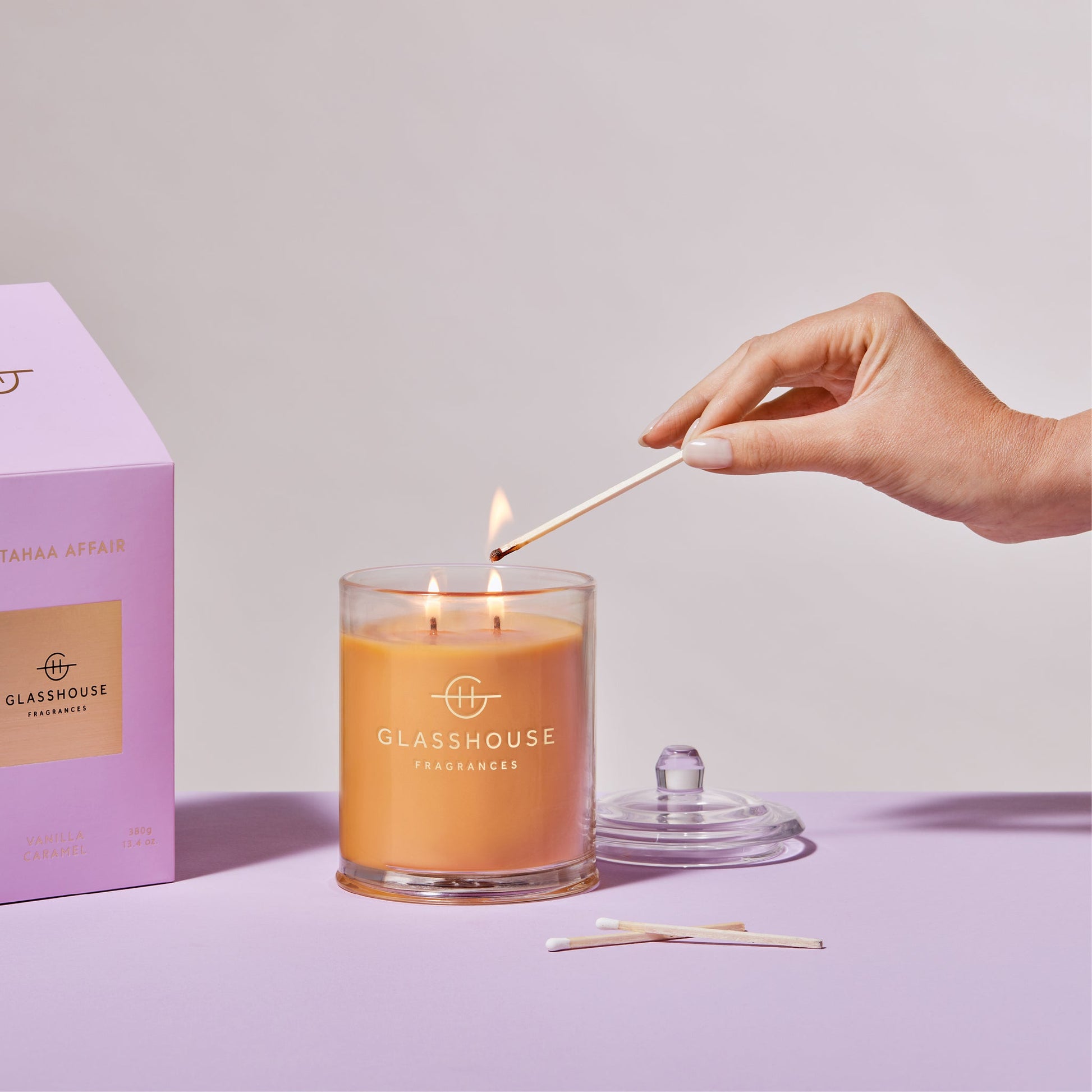 a womans hand lighting the A Tahaa Affair Triple Scented Candle while displayed next to the pink box