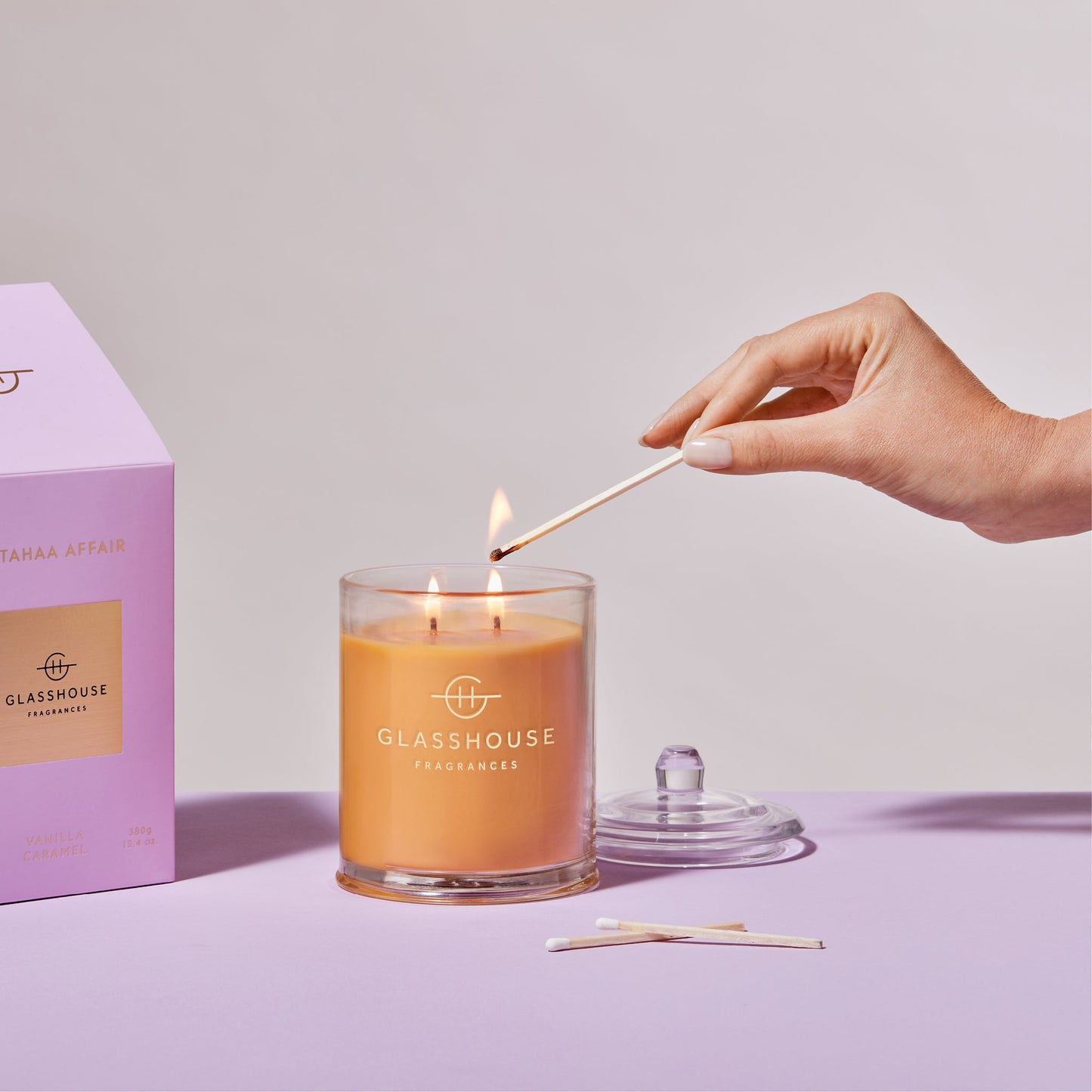 a person's hand lighting the A Tahaa Affair Triple Scented Candle while displayed next to the pink box