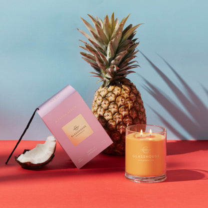 A Tahaa Affair Triple Scented Candle jar displayed next to the pink box, chunk of coconut and whole pineapple on a corral surface