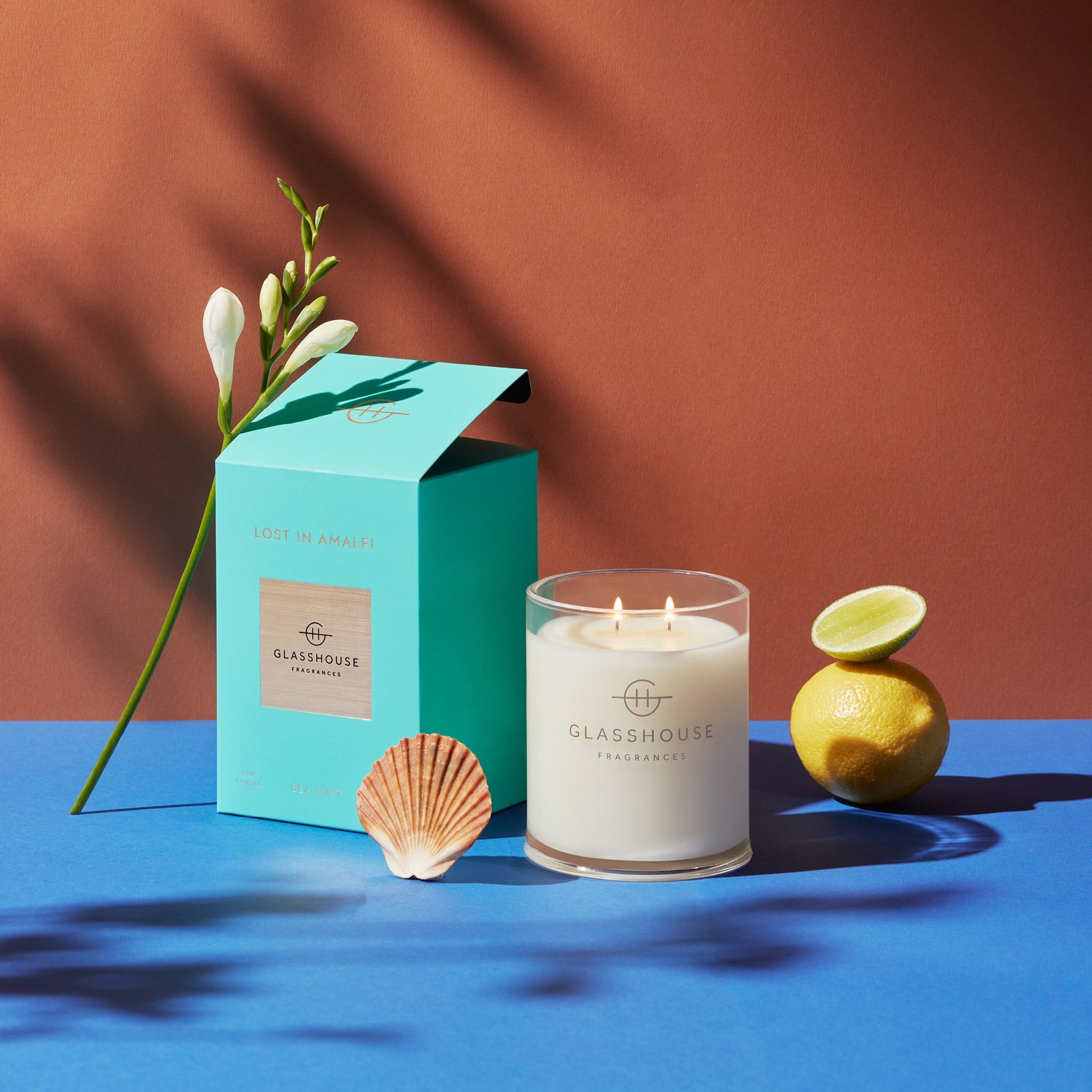 Lost In Amalfi Triple Scented Candle jar displayed next to the turquoise box, lemons, sea shell, and sprig of flowers against a blue and mauve background