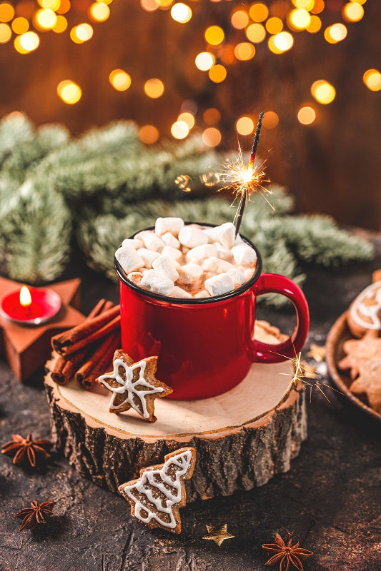 cup mug filled with cocoa and topped with marshmallows set on a slab of wood surrounded by greenery, spices, and cookies.