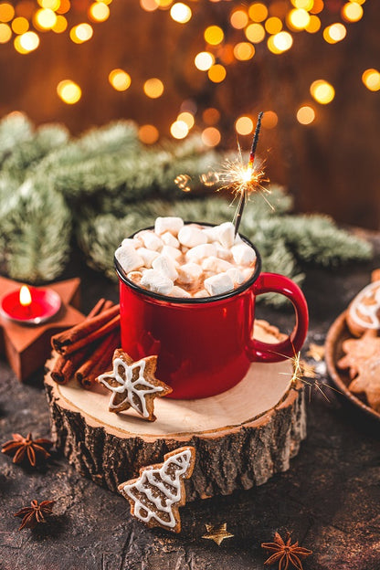 red mug filled with hot chocolate topped with marshmallows set on a slab of wood on a table surrounded by greenery, cookies, and spices.