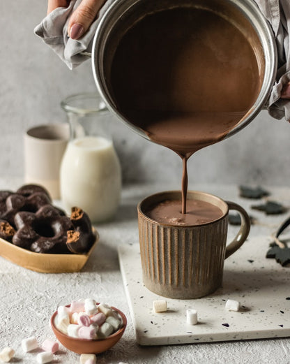 hands holding pot poring sugar free hot chocolate into a mug on a table with chocolate bits and marshmallows scattered around.