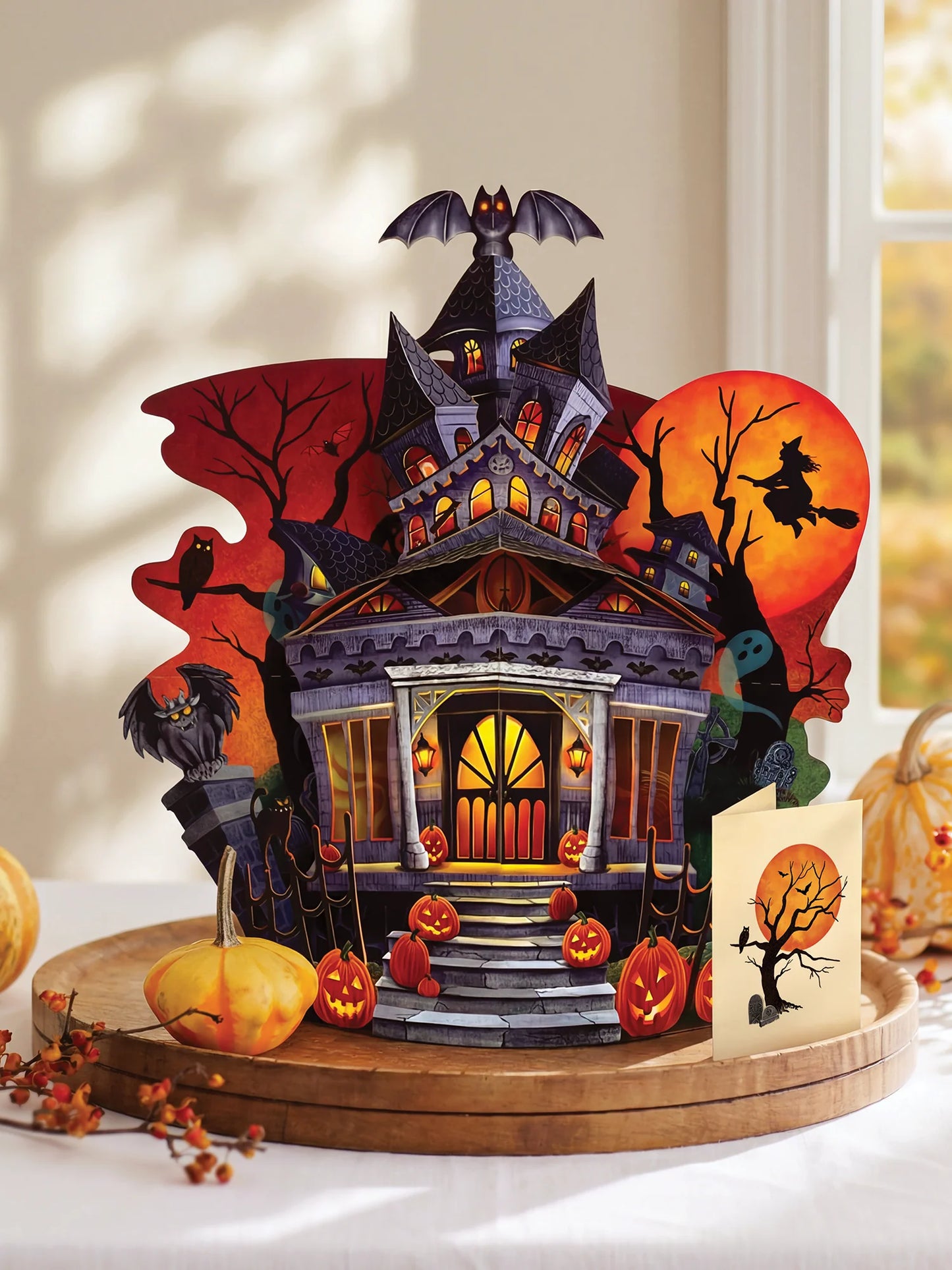 paper pop up haunted house card featuring a haunted house, bats, pumpkins, and a witch silhouette against orange moon and sun shapes, displayed on a wooden tray with a matching greeting card in a bright indoor setting.