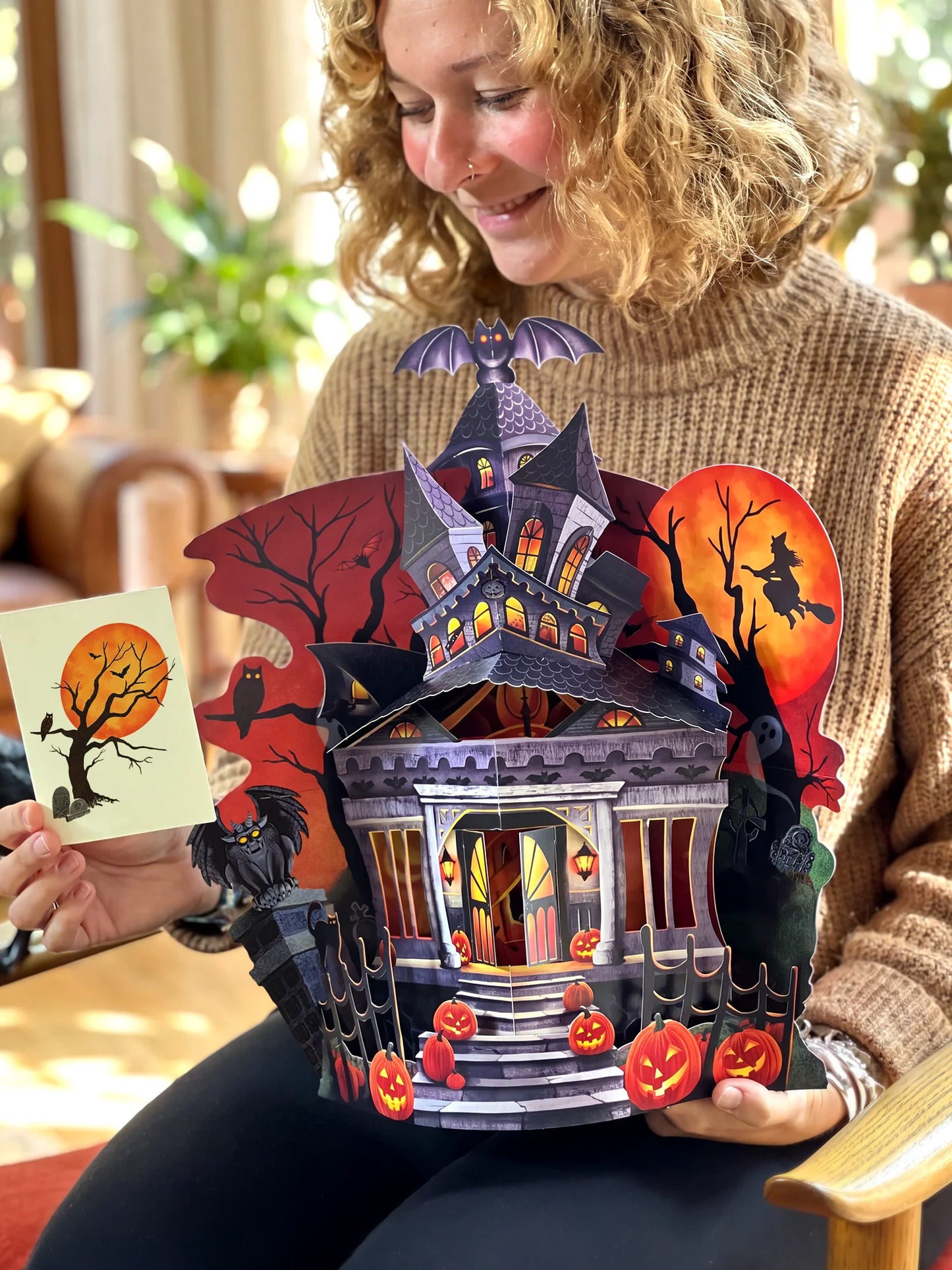 person holds a large, multi-layered 3D Halloween pop-up paper house. The pop-up features a spooky haunted mansion, jack-o'-lanterns, a witch silhouette on a full moon, and a black tree.