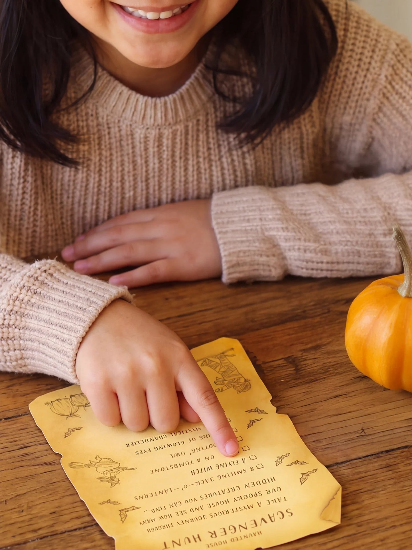 A person in a beige knit sweater points to a yellow parchment-style scavenger hurt card on a wooden table, with a small orange pumpkin nearby.