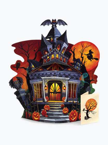 haunted house pop up card with jack-o'-lanterns, bats, a witch on a broom against a large orange moon, and a small greeting card in front on a light gray background.
