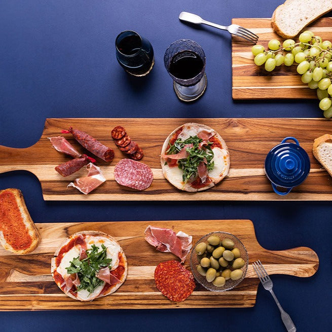 3 table planks with a variety of meats, olives, and grapes on them arranged on a blue background with glasses of wine.