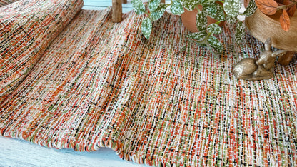A close-up of a woven textile runner with a multicolor blend of orange, red, green, beige, and grey tarns, styled with a potted plant and a small gold deer figurine.