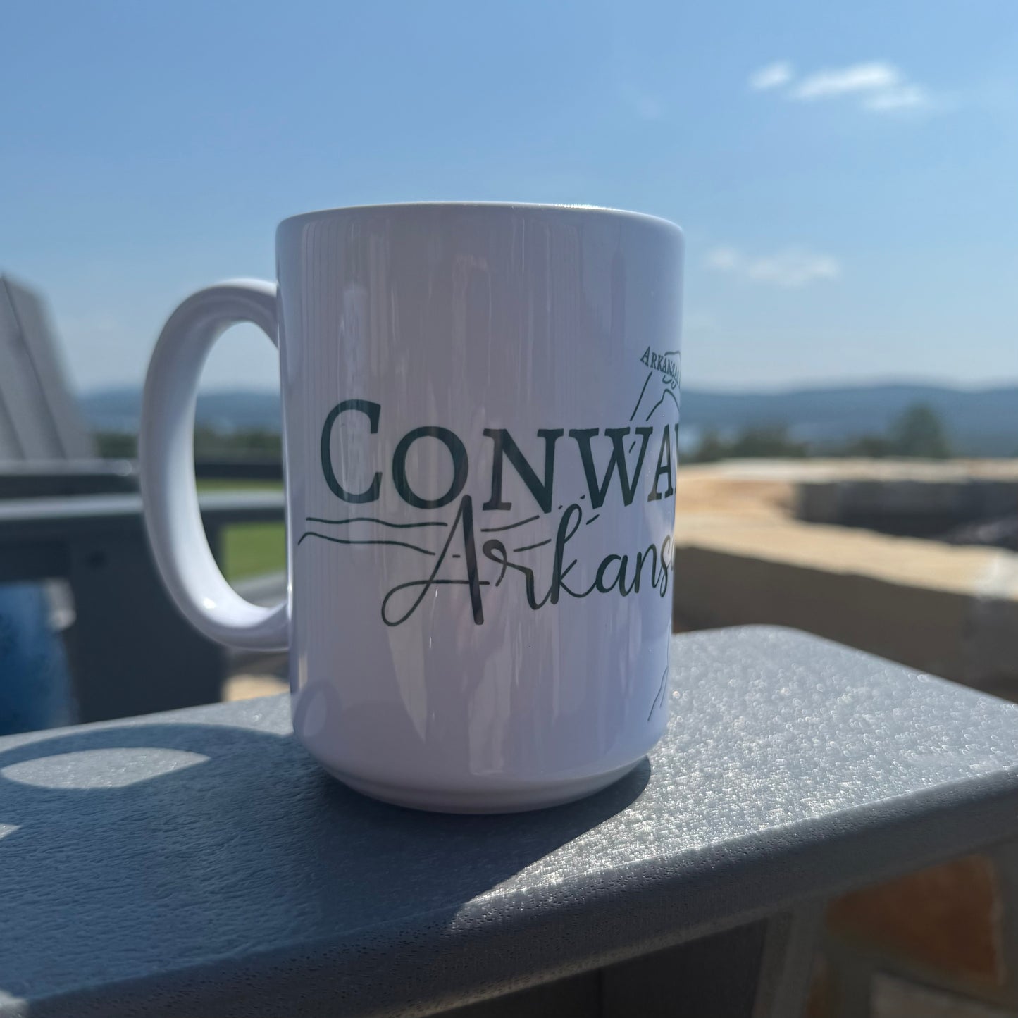 White mug with 'Conway Arkansas' text on a patio chair armrest with a scenic Arkansas view on the background