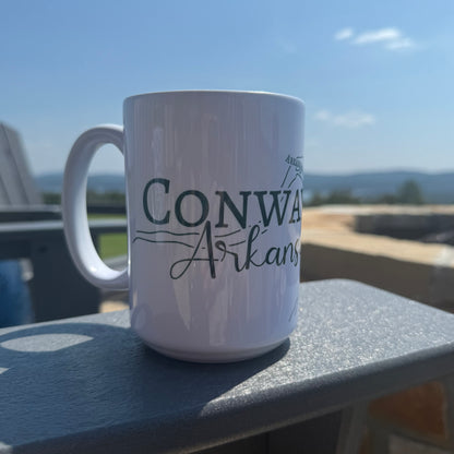 White mug with 'Conway Arkansas' text on a patio chair armrest with a scenic Arkansas view on the background
