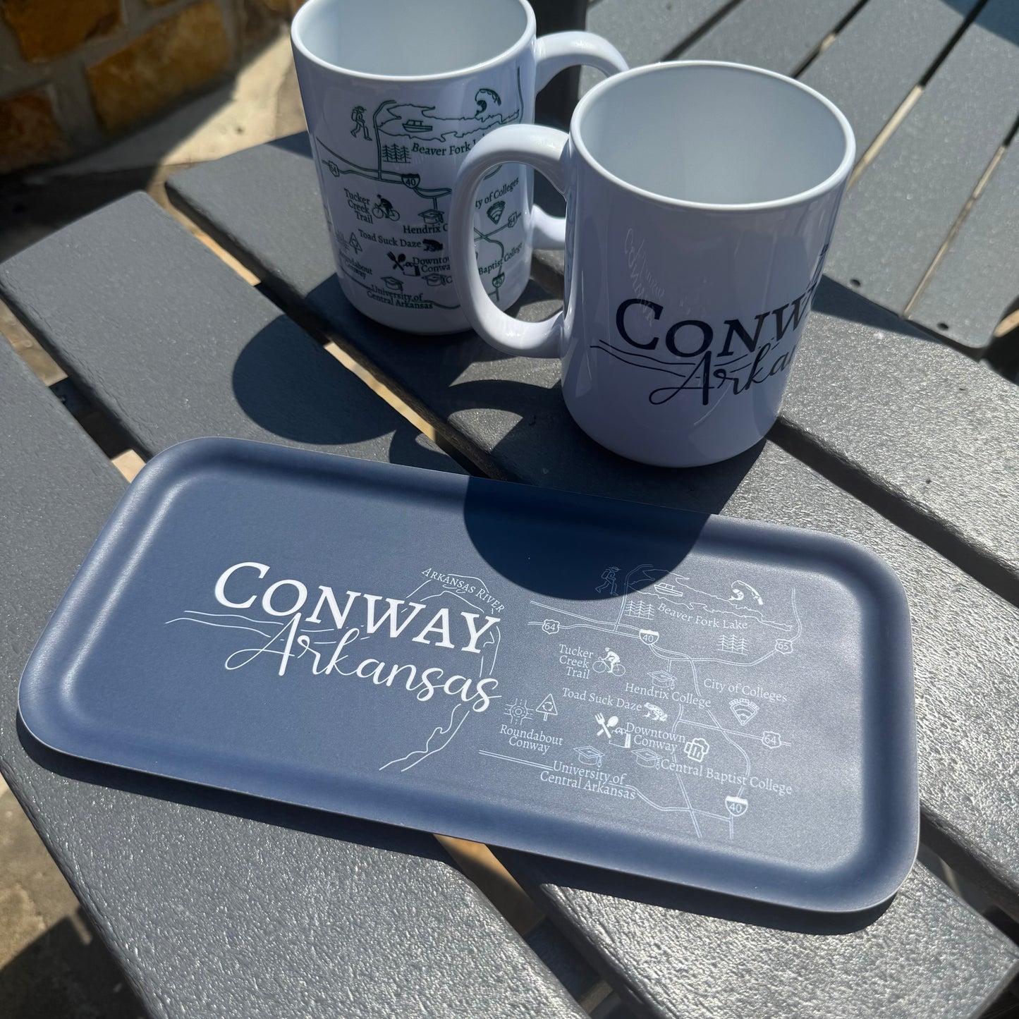 Two mugs and a tray with 'Conway Arkansas' design on a wooden patio table.