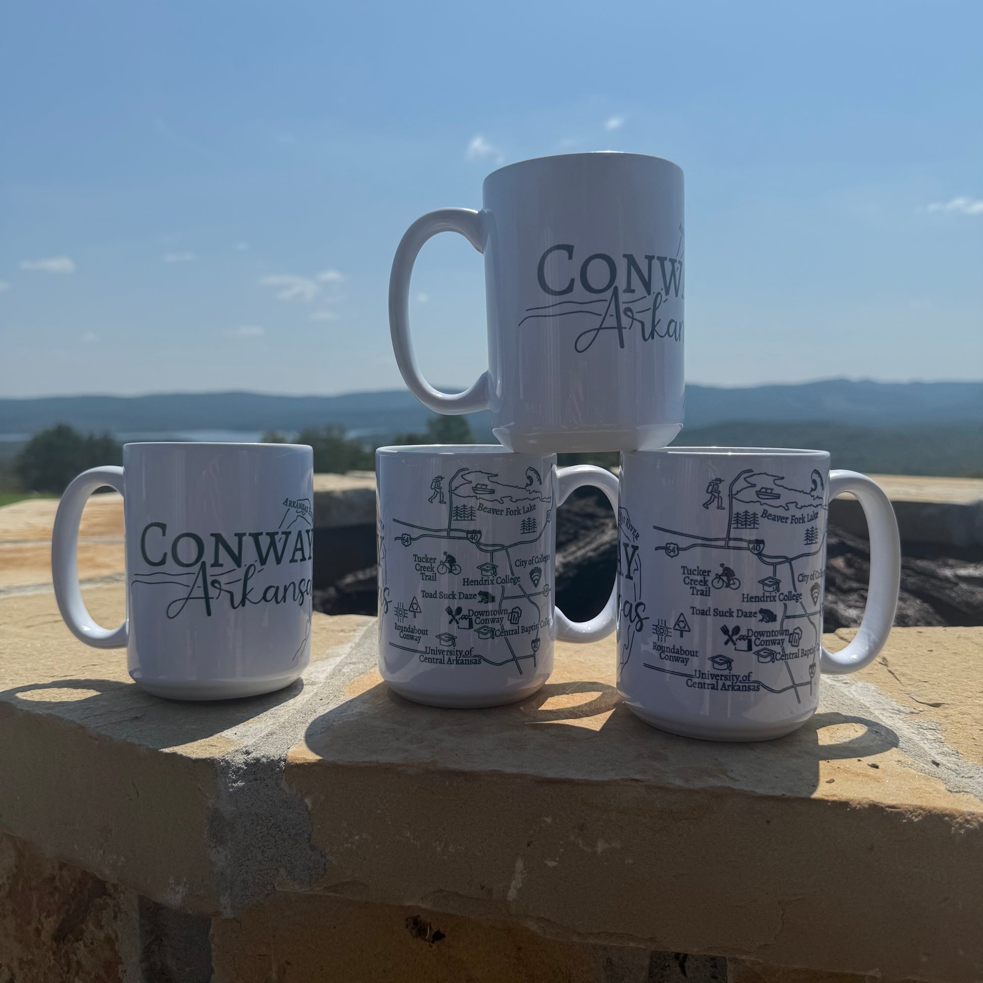 Four white mugs with 'Conway Arkansas' text on a stone ledge with a scenic Arkansas background.