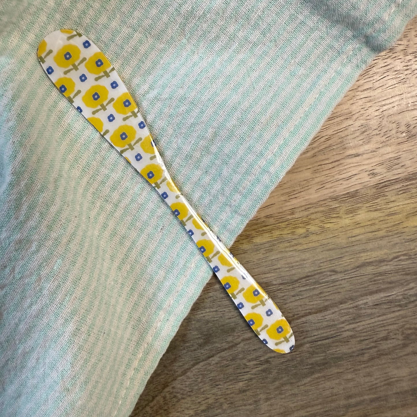 white spreader with yellow floral pattern laying on a wooden surface with green striped towel