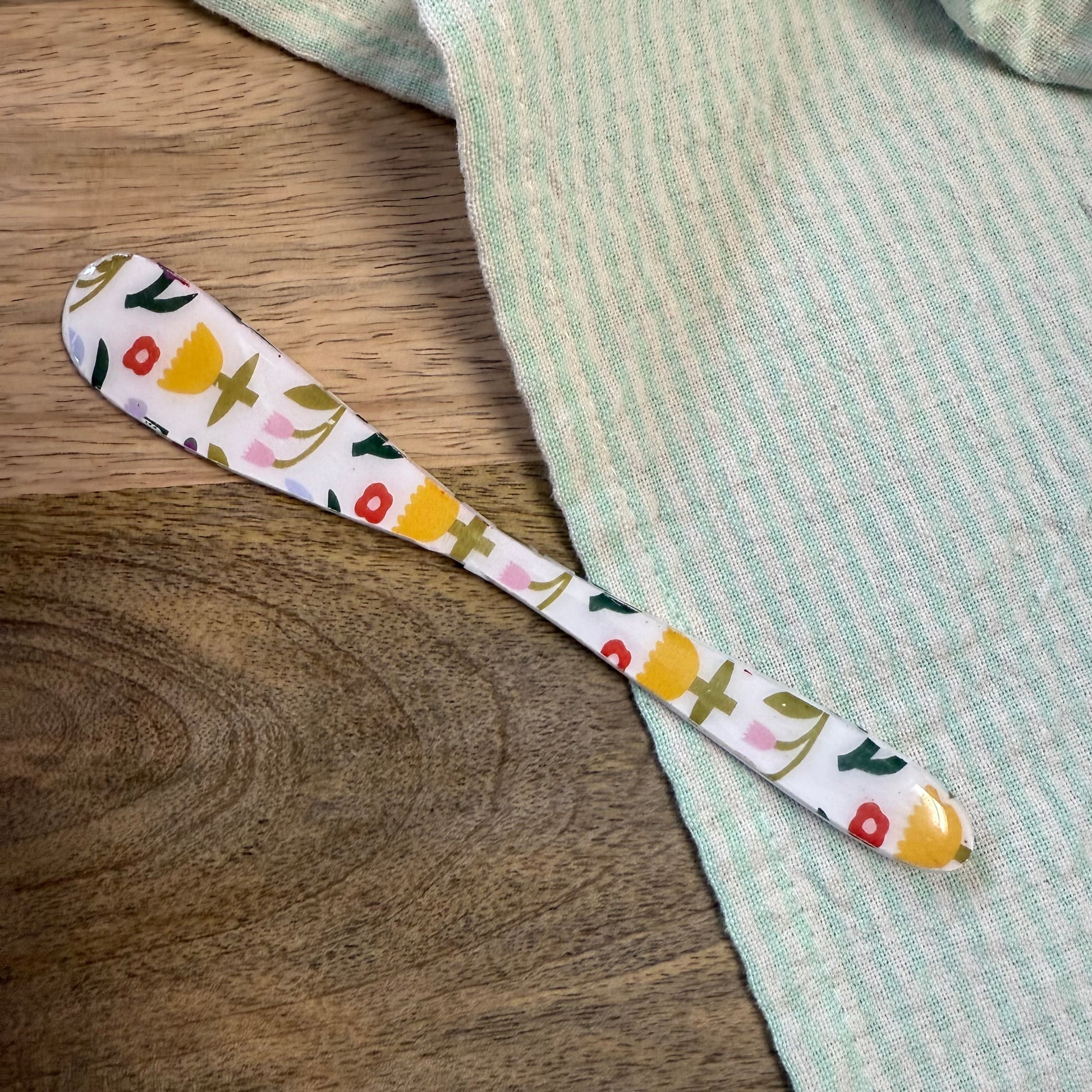 white spreader with multi-color floral pattern laying on a wooden surface with green striped towel