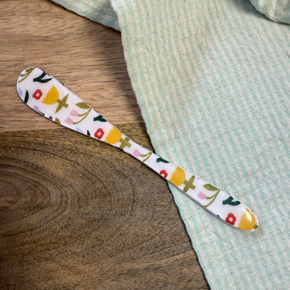 white spreader with multi-color floral pattern laying on a wooden surface with green striped towel