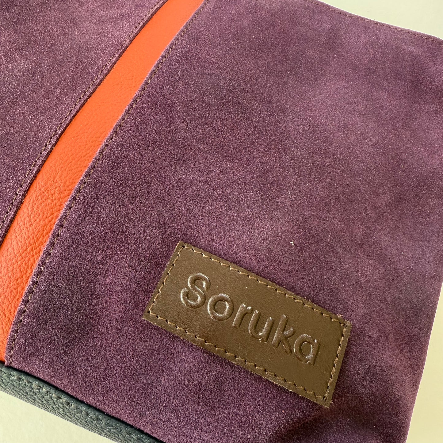 Close-up of a purple suede bag with a dark orange zipper and 'Soruka' brand label.