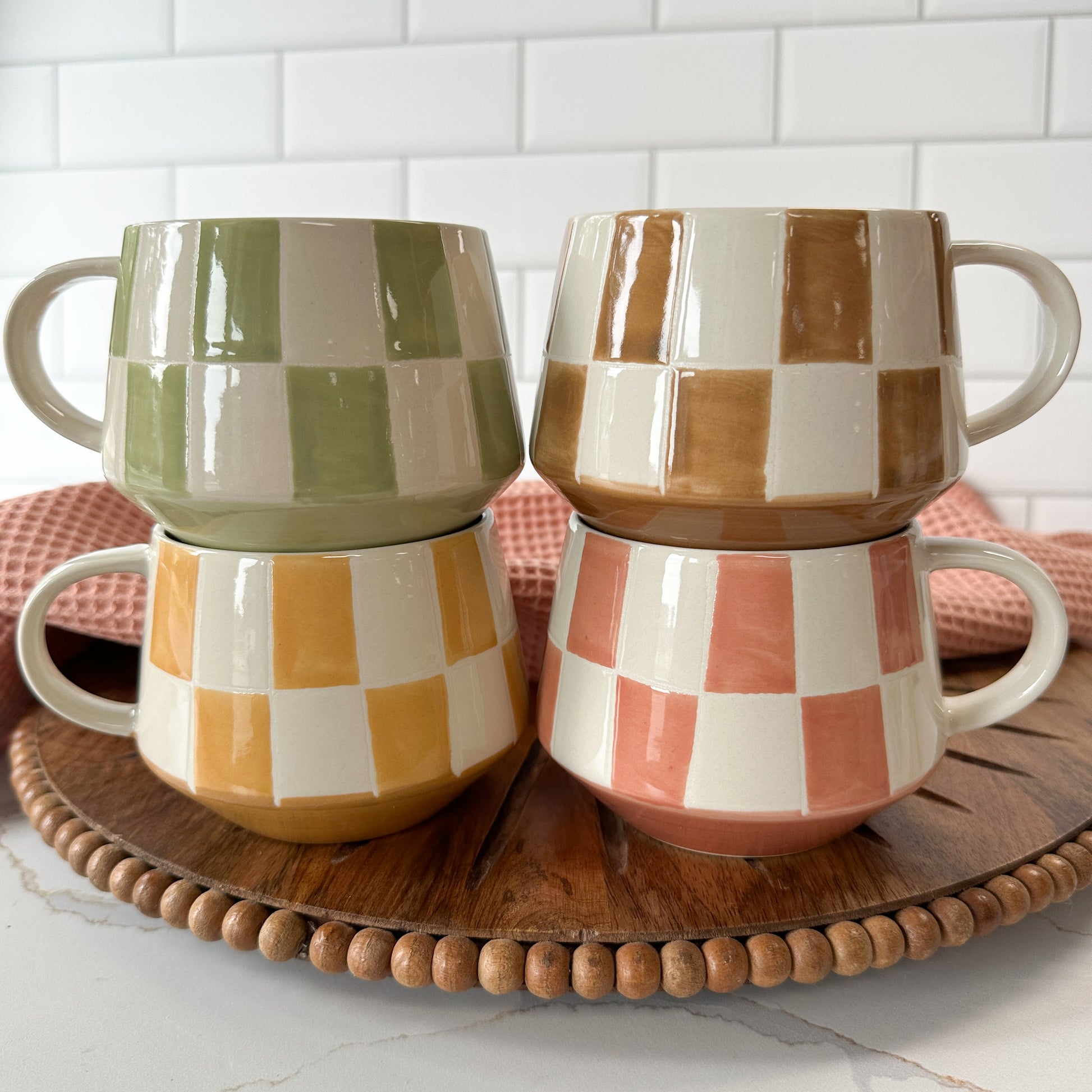 Four checkered ceramic mugs stacked on a wooden tray with a white tiled wall background.