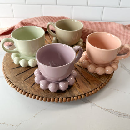 4 colors of Stoneware Mugs with Bubble Saucers set on a round wooden board with a blush dishtowel in the background